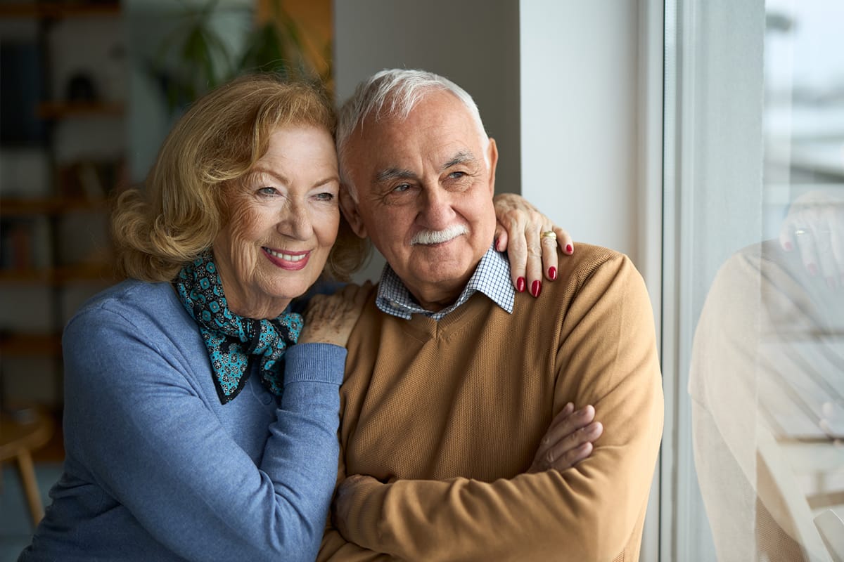 Pareja de adultos mayores abrazados y sonrientes mirando juntos por la ventana.