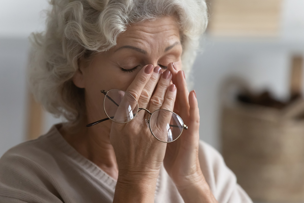 Mujer mayor frotándose los ojos mientras sostiene sus lentes, lo que sugiere fatiga o molestia visual.