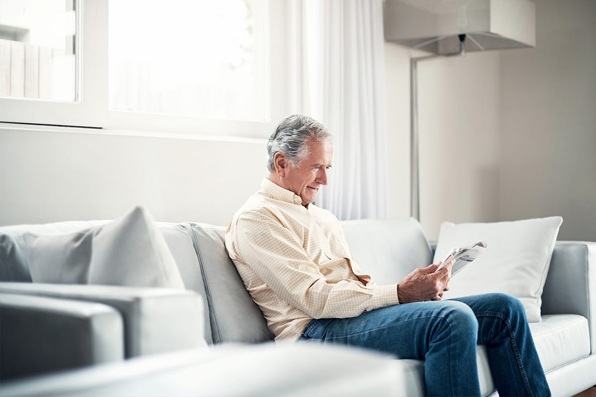 Hombre mayor sentado en un sofá en casa, leyendo el periódico en una sala luminosa.