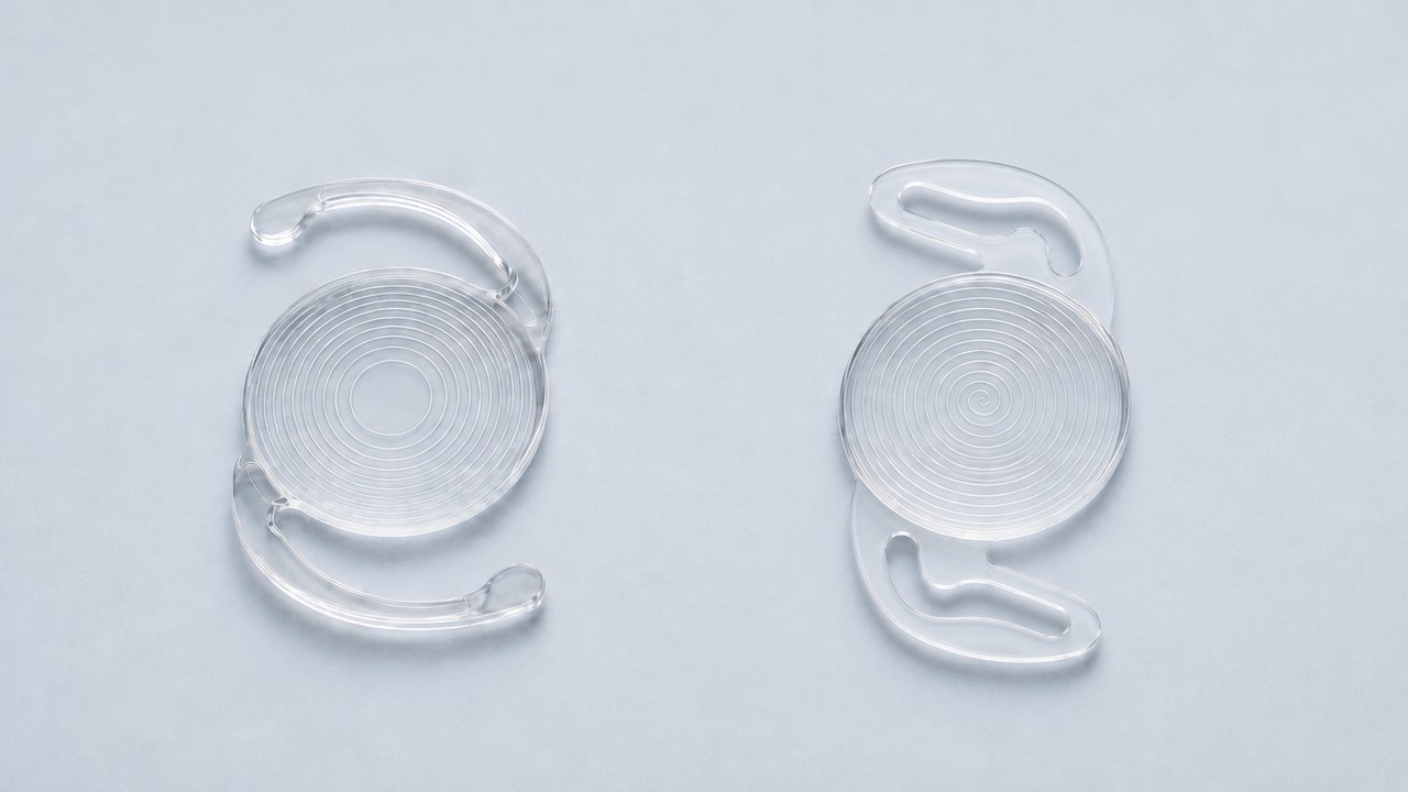 Rendering of two clear lens implants: the left lens has separate closed concentric rings and thin curved support arms, and the right lens has one continuous groove that spirals gradually outward like the groove on a record, with the broad closed-loop support arms of the Rayner lens